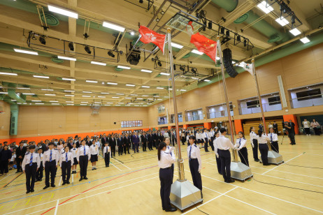 Guests and students stand solemnly, gazing attentively at the flag
